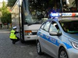 policyjny radiowóz, a za nim autobus. Przy przednim prawym kole autobusu jest policjant w pozycji kucającej i sprawdza stan techniczny pojazdu