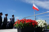 moment unoszenia flagi państwowej. Stojący po lewej stronie policjanci patrzą na masz z flagą. na pierwszym planie kwoaty w kolorze biało-czerwonym