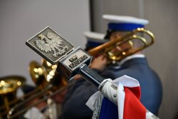 godło Polski Orzeł Biały na metalowym zakończeniu drzewca flagi, w tle członek policyjnej orkiestry