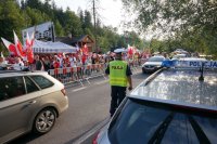 po prawej stronie radiowóz, za nim na drodze znajduję się policjant, który obserwuje tłum kibiców w biało czerwonych barwach, którzy idą lewą stroną drogi