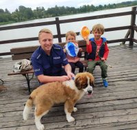 policjant w półprzysiadzie oraz siedzące za nim dzieci. Widać niebo, wodę i pomost na którym znajdują się osoby na fotografii. Przed policjantem mały pies