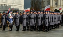 policjanci stoją w kolumnach,  tle powiewają flagi Polski