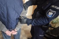 policjant trzyma zatrzymanego za ramię, zatrzymany kajdanki na rękach z tyłu