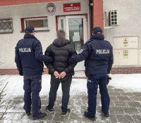 policjanci stoją przed budynkiem, dzień,śnieg pomiędzy nimi mężczyzna z kajdankami na rękach z tyłu, policjanci trzymają go za ramię