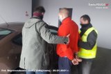 Policjanci trzymają zatrzymanego mężczyznę za ręce, które ma spięte z tyłu kajdankami. Wszyscy stoją obok nieoznakowanego radiowozu