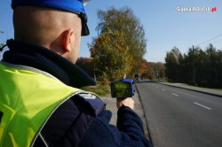 grafika- policjant mierzy prędkość, stoi obok drogi, kieruje miernik w stronę poruszających się samochodów, dziń