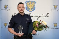dzielnicowy mł.asp. Kamil Przewieźlik trzyma statuetkę oraz kwiaty, jest w mundurze, na tle baneru Starostwa Powiatowego w Cieszynie