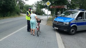 grafika-policjant rozmawia z kobieta prowadzącą rower, po prawej stronie radiowóz policji