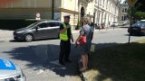 dzień, policjant rozmawia z pieszymi na drodze, w tle budynek i samochód