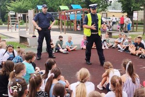 grafika-dzień, park,plac zabaw,policjant przemawia do grupy dzieci