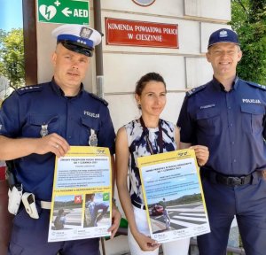 grafika-dzień, słońce, zdjęcie w plenerze, osoby trzymają plakaty i pozują do zdjęcia przed budynkiem Komendy Powiatowej Policji w Cieszynie