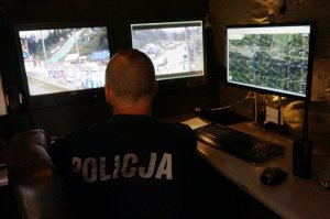 policjant odwrócony tyłem do zdjęcia, patrzy na ekrany monitoringu,ciemno, pomieszczenie dowodzenia, napis policja na koszulce