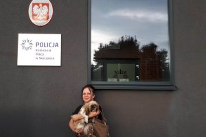 właścicielka psa trzymie go na rękach, stoi przed budynkiem komisariatu policji w Skoczowie