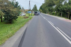 Na asfaltowej drodze widoczne ślady hamowania. Dalej stoi uszkodzony samochód w polu. Po prawej na drodze stoi policyjny samochód służbowy