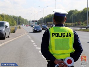 policjant na drodze obserwuje ruch pojazdów, z tyłu trzyma tarczę do zatrzymywania pojazdów