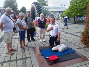 dzień, rynek, policjant w mundurze klęczy przy fantomie do resuscytacji i tłumaczy zebranym wokoło dzieciom oraz dorosłym