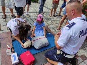 dzień, rynek, policjant w mundurze klęczy przy fantomie do resuscytacji i tłumaczy zebranym wokoło dzieciom oraz dorosłym