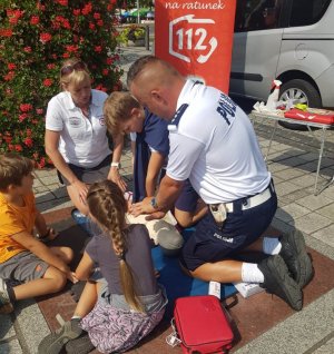 dzień, rynek, policjant w mundurze klęczy przy fantomie do resuscytacji i tłumaczy zebranym wokoło dzieciom oraz dorosłym