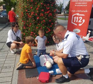 dzień, rynek, policjant w mundurze klęczy przy fantomie do resuscytacji i tłumaczy zebranym wokoło dzieciom oraz dorosłym