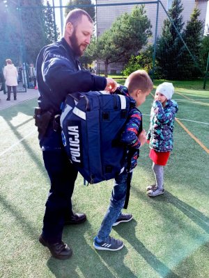 grafika,dzień, policjant w mundurze zakłada dziecku plecak na plecy z napisem policja