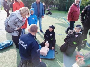 grafika, dzień, boisko sportowe, słońce, policjant klęczy i demonstruje RKO na manekinie, przyglądają się mu inne osoby