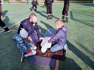 grafika, dzień, boisko sportowe, słońce, policjant klęczy i demonstruje RKO na manekinie, przyglądają się mu inne osoby