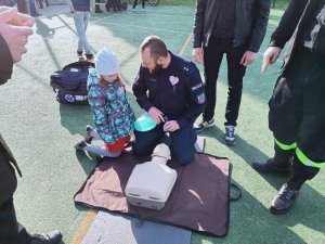 grafika, dzień, boisko sportowe, słońce, policjant klęczy i demonstruje RKO na manekinie, przyglądają się mu inne osoby