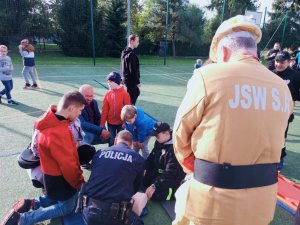 grafika, dzień, boisko sportowe, słońce, policjant klęczy i demonstruje RKO na manekinie, przyglądają się mu inne osoby