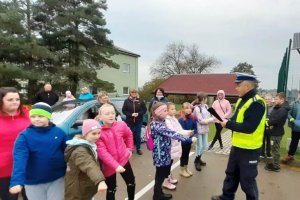 grafika-dzień, budynek OSP w tle, ludzie zebrani wokół policjanta odbierają odblaski