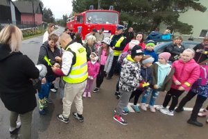 grafika-dzień, samochód straży pożarnej w tle, ludzie zebrani wokół osoby z napisem MEDIA na plecach odbierają odblaski