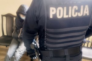 grafika-dzień, wewnątrz budynku, policjant stoi odwrócony plecami, na mundureze napis Policja, w tle widać siedzącego mężczyznę z  kapturem na głowie