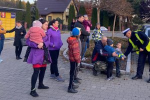 grafika-dzień, poza budynkiem policjant wręcza odblaski licznie zgromadzonym ludziom