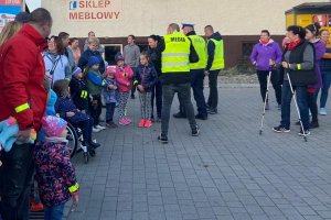 grafika-dzień, poza budynkiem policjant wręcza odblaski licznie zgromadzonym ludziom