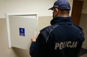 grafika- policjant stoi tyłem i sprawdza skrzynkę z bezpiecznikami prądu