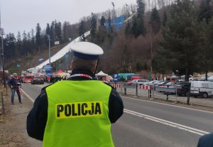 grafika-dzień, policjant stoi przy drodze, kieruje ruchem, w tle skocznia