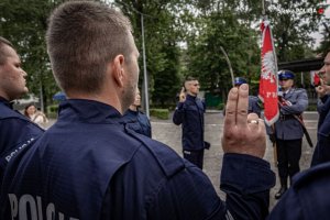 grafika- dzień, otwarta przestrzeń , policjanci ślubują, prawa ręka uniesiona, w tle sztandar