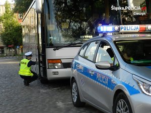 grafika, dzień, policyjny radiowóz, w tle policjant w przysiadzie sprawdza koło autobusu