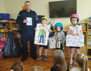 grafika-policjant z dziećmi przedszkola pozuje do zdjęcia, dzieci założone na głowie kaski-nagrody z konkursu