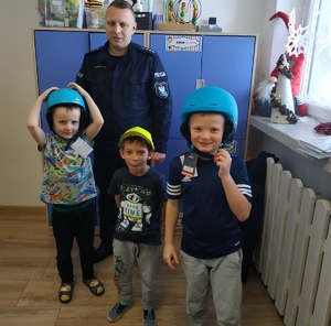 grafika-policjant z dziećmi przedszkola pozuje do zdjęcia, dzieci założone na głowie kaski-nagrody z konkursu