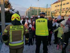 grafika-dzień, śnieg, poza budynkiem, policjant rozmawia z liczną grupą ludzi i wręcza im odblaski