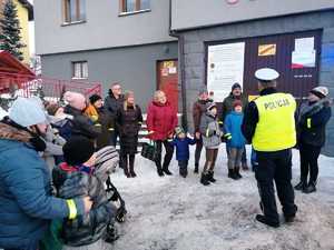 grafika-dzień, śnieg, poza budynkiem, policjant rozmawia z liczną grupą ludzi i wręcza im odblaski