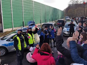 grafika- dzień, policjanci stoją przed radiowozem, policjanci pozują do zdjęcia z maskotką, ludzie robią im zdjęcia