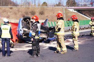 grafika- dzień, błękitne niebo, jezdnia,policjanici strażacy na miejscu wypadku drogowego przy uszkodzonym samochodzie