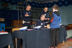 grafika- policjanci za stolikiem sędziowskim, sala gimnastyczna, uczeń stoi  i zgłasza swoje dane