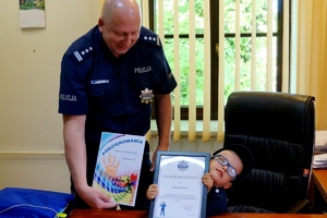 grafika-pomieszczenie, Szymonek siedzi na fotelu komendanta za biurkiem, a Komendant insp.Jacek Stelmach stoi obok, oboje prezentują otrzymane prezenty