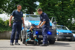 grafika-plac , słoneczna pogoda, policjant nakłada czapkę Szymkowi, który siedzi na wózku inwalidzkim, w tle radiowóz