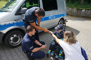 grafika-plac , słoneczna pogoda, policjanci pozują do zdjęcia razem z mamą Szymka i dziękują mu za wizytę