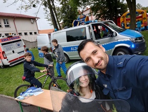 grafika-dzień, park ,policjant pozuje do zdjęcia  z dzieckiem ubranym w policyjny kask, w tle radiowóz