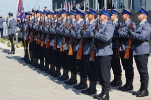 grafika, dzień, plac, policjanci w mundurach galowych stoją w szeregu i trzymają reprezentacyjnie broń długą