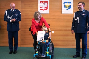 grafika-sala, dziecko na wózku inwalidzkim, chłopiec trzyma w ręku akt mianowania,komendanci stoją po lewej i prawej stronie